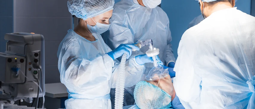 OP-Team bei Beatmungsmanagement: Fachkräfte in Schutzkleidung bereiten Beatmungsmaske und -schlauch für narkotisierten Patienten vor. ATAs überwachen Vitalfunktionen und sichern Atemwege während Operationen.