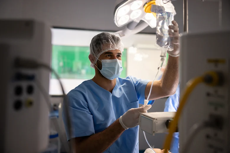 Anästhesietechnischer Assistent im OP: Medizinischer Fachmann mit Haube, Maske und Handschuhen überprüft Infusionsbeutel vor einer Operation. ATA unterstützen bei Narkosevorbereitung und Patientenüberwachung.