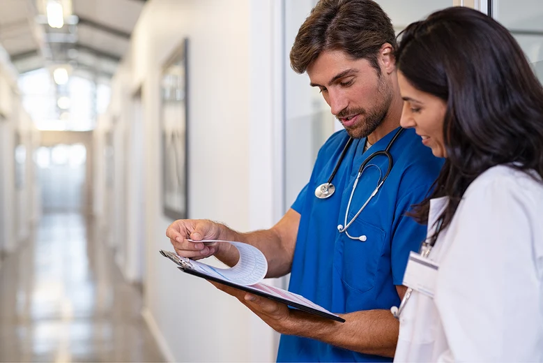 Assistenzarzt und Kollegin besprechen Patientenakte auf Klinikflur. Assistenzärzte absolvieren nach dem Medizinstudium eine 5-6-jährige Facharztweiterbildung unter ärztlicher Supervision in Kliniken.