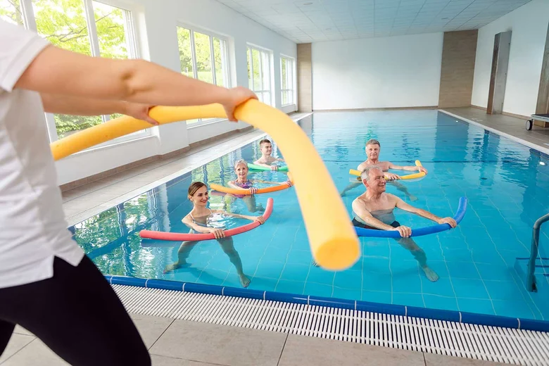Aquafitness-Kurs im Hallenbad: Trainer/in leitet Gruppe mit bunten Schwimmnudeln bei Wassergymnastik. Fachangestellte für Bäderbetriebe organisieren Schwimmkurse, überwachen Sicherheit und Wasserqualität.