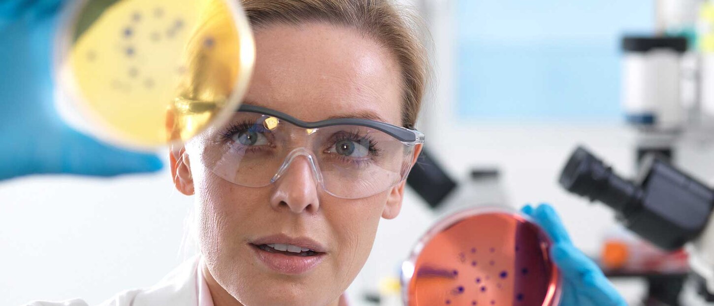 Eine Laborantin mit Schutzbrille blickt in eine Kultur in einer Petrischale.