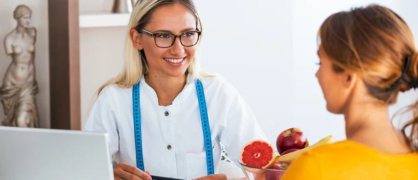 Diätassistentin berät Patientin mit Maßband und frischem Obst am Laptop. Diätassistenten erstellen individuelle Ernährungspläne, berechnen Nährstoffbedarf und schulen bei therapeutischer Ernährungsumstellung.