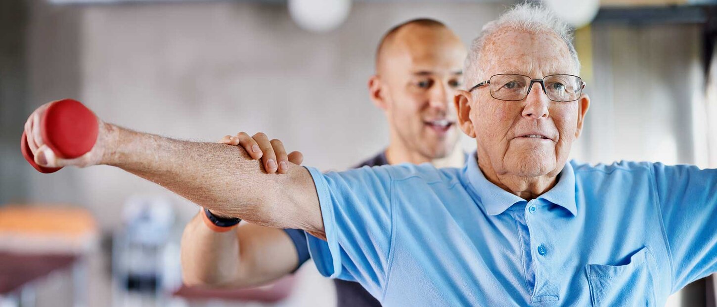 Ein Ergotherapeut steht hinter einem älteren Mann, der eine Hantel in der Hand hält und die Arme zur Seite ausstreckt. 