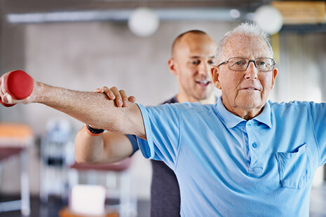 Ein Ergotherapeut steht hinter einem älteren Mann, der eine Hantel in der Hand hält und die Arme zur Seite ausstreckt.