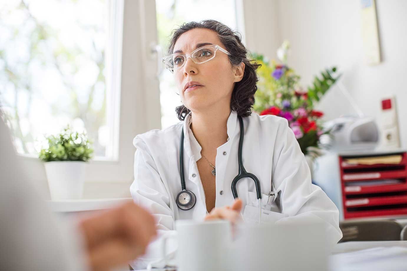 Eine Ärztin mittleren Alters und Brille blickt konzentriert auf ihren Patienten. Sie sitzt ihm gegenüber an einem Schreibtisch.