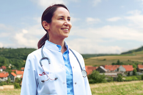 Eine Ärztin (Frau im weißen Kittel und mit Stetoskop um den Hals) steht vor einer Wiese. Im Hintergrund ist ein Dorf zu sehen.