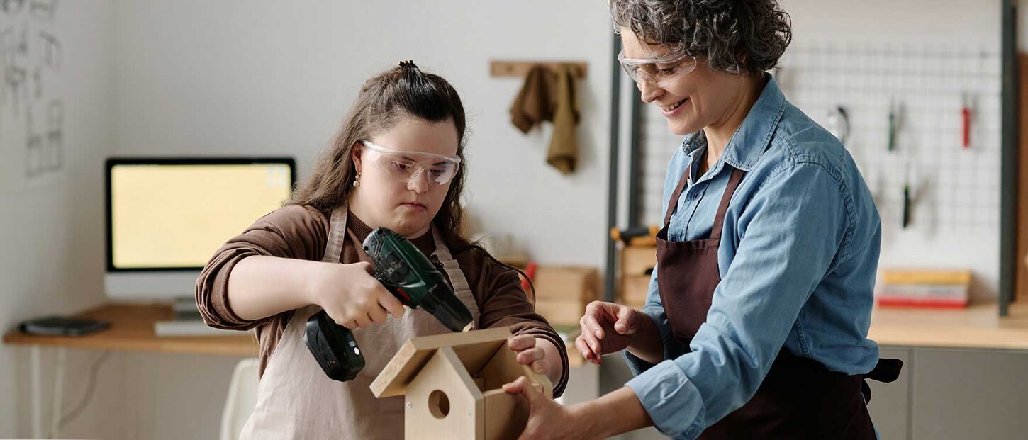 Heilerziehungspflegerin baut mit Dame mit Handcap ein Vogelhaus aus Holz. Sie stehen in einer Holzwerkstatt, haben Sicherheitsbrillen auf und Werkzeuge in den Händen.