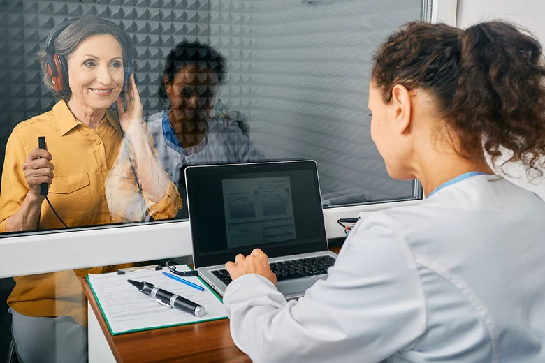 Hörgeräteakustikerin führt computergestützten Hörtest in Schallkabine durch: Kundin trägt Kopfhörer, Messergebnisse werden am Laptop erfasst. Professionelle Audiometrie ermittelt Hörschwellen und bildet Grundlage für individuelle Hörgeräte-Anpassung und V