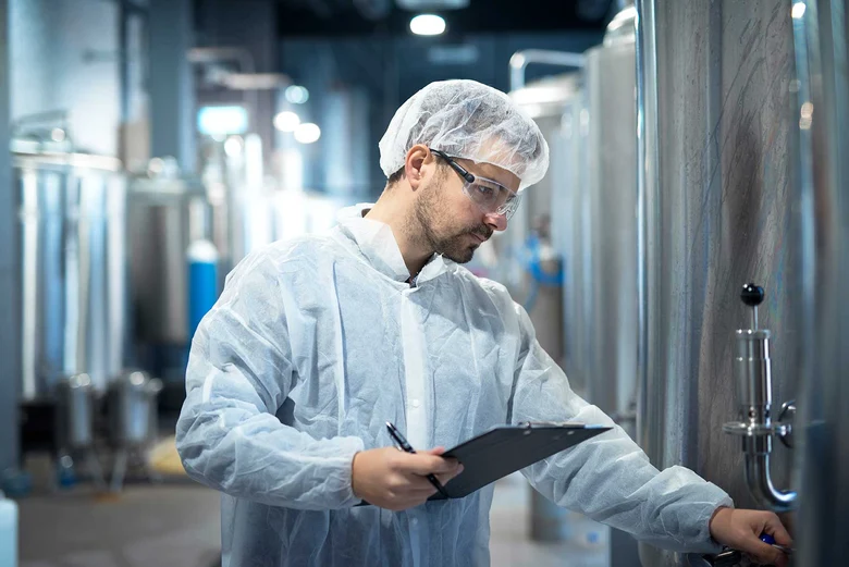 Hygienekontrolleur in Schutzkleidung mit Haarhaube dokumentiert Kontrolle in Lebensmittelproduktion am Klemmbrett. Überwacht Hygienestandards, Produktionsanlagen und Reinigungsprozesse in der Industrie zum Schutz der Verbrauchergesundheit.