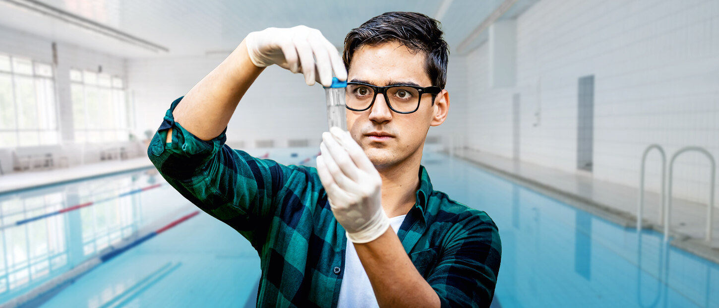 EIn männlicher Hygienekontrolleur steht in einem Schwimmbad und guckt in ein Reagenzglas mit Wasserprobe.