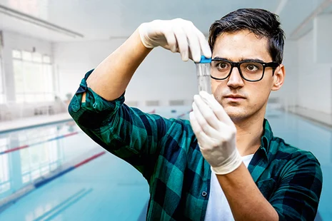 Hygienekontrolleur entnimmt Wasserprobe im Schwimmbad mit Schutzhandschuhen und Reagenzglas. Kontrolliert Wasserqualität, pH-Wert, Chlorgehalt und mikrobiologische Parameter zum Schutz vor Keimen und Krankheitserregern in öffentlichen Bädern.