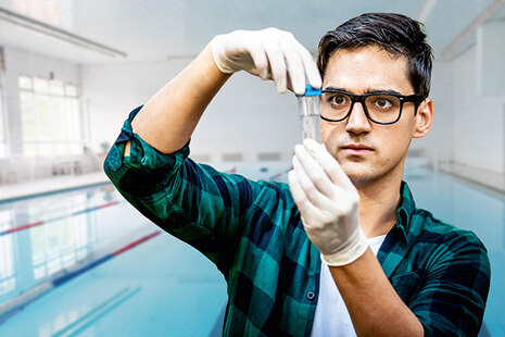 Ein junger Mann in einem weißen Kittel und mit Mundschutz steht in einem Labor und entnimmt mit einer Pipette etwas aus einer Petzrischale.
