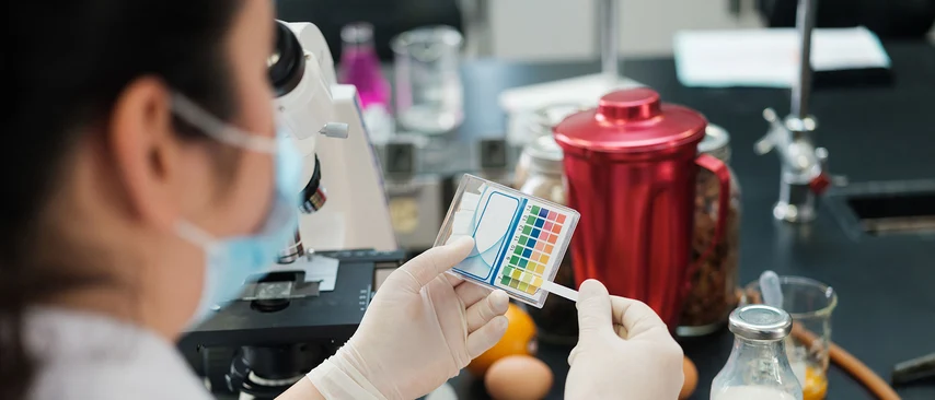 Lebensmittelchemikerin analysiert mit Farbskala Milchproben im Labor mit Mikroskop. Lebensmittelchemiker untersuchen Lebensmittel auf Qualität, Inhaltsstoffe, Schadstoffe und Kennzeichnung zum Schutz der Verbraucher und Einhaltung gesetzlicher Vorgaben.