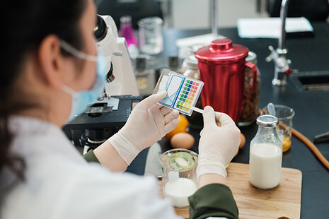 Eine Lebensmittelchemikerin prüft mittels Teststreifen den Wert eines Lebensmittels. Sie hält das Teststäbchen an eine Auswertungstabelle.