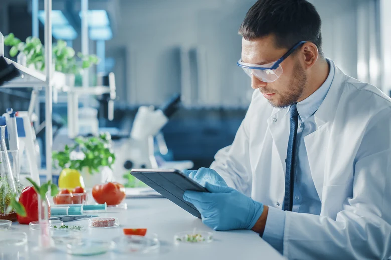 Lebensmitteltechniker mit Schutzbrille prüft Gemüseproben und Tablet im Labor mit Pflanzen. Lebensmitteltechniker entwickeln, optimieren und überwachen Herstellungsprozesse, kontrollieren Qualität und Hygiene bei der industriellen Lebensmittelproduktion.