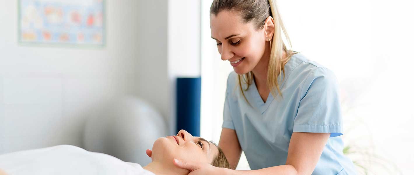 Eine Masseurin beugt sich über eine liegende Patientin, greift an ihren Nacken und lächelt sie an.