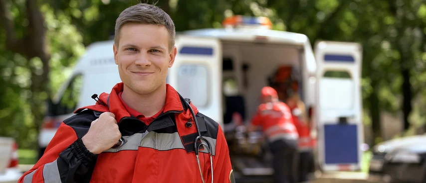 Notfallsanitäter in Einsatzkleidung vor Rettungswagen im Außeneinsatz. Notfallsanitäter sind erste medizinische Hilfe am Notfallort, führen lebensrettende Maßnahmen durch und stabilisieren Patienten bis zur Klinikeinlieferung in kritischen Situationen.