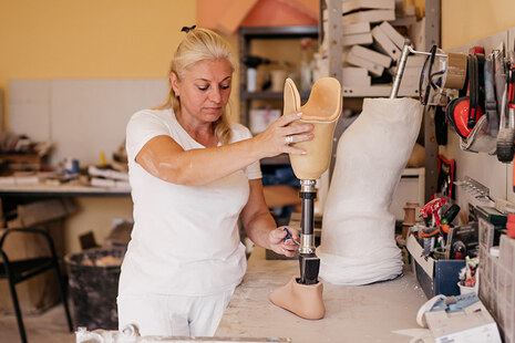 Frau steht in einer Werkstatt. Sie hat auf dem Tisch vor sich eine Beinprothese, an der sie schraubt.