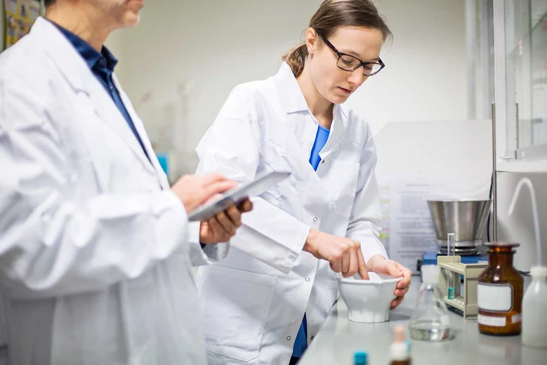 Pharmazeutisch-technische Assistentin im Laborkittel bei der Arzneimittelherstellung, arbeitet mit Mörser und Dokumenten im Apothekenlabor.
