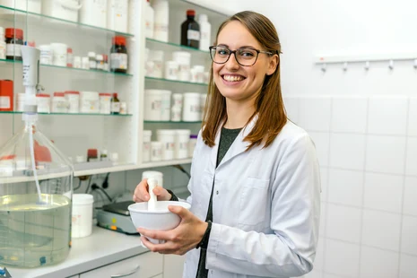 Pharmazeutisch-technische Assistentin im Laborkittel mit Mörser und Stößel in einer Apotheke, im Hintergrund Arzneimittelregale.