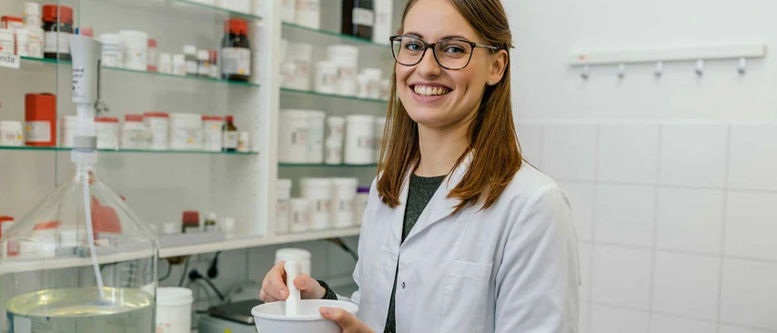 Pharmazeutisch-technische Assistentin im Laborkittel mit Mörser und Stößel in einer Apotheke, im Hintergrund Arzneimittelregale.