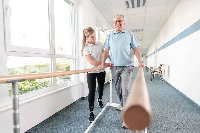 Physiotherapeutin unterstützt älteren Mann beim Gangtraining am Gehbarren in einer Rehabilitationseinrichtung.