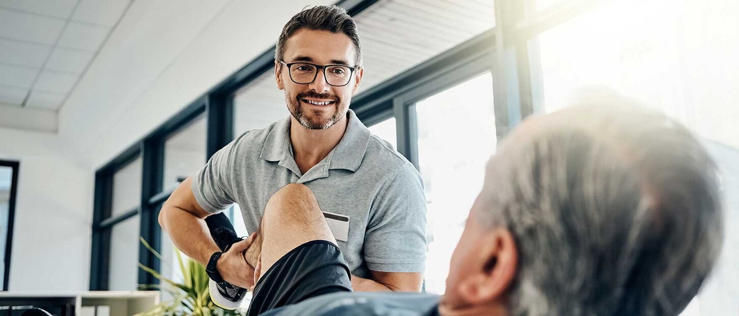 Physiotherapeut beugt das Knie seines liegenden Patienten und guckt lächelt diesen an.