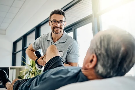 Physiotherapeut behandelt Bein eines Patienten in Praxisraum, hält den Unterschenkel und führt eine Bewegungsübung durch.