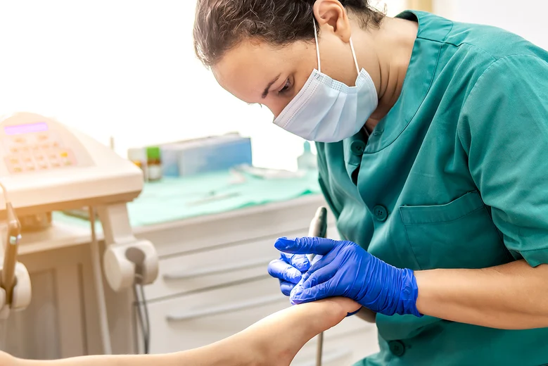 Podologin mit Mundschutz und Handschuhen untersucht Fuß eines Patienten in Behandlungsraum.