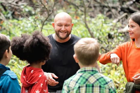 Sozialpädagoge im Gespräch mit vier Kindern im Freien. Natürliche Umgebung mit Bäumen im Hintergrund.