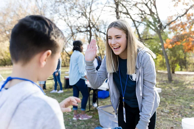 Sozialpädagogin mit Schlüsselband gibt Jugendlichen im Park High Five. Weitere Personen im Hintergrund bei Gruppenaktivität.