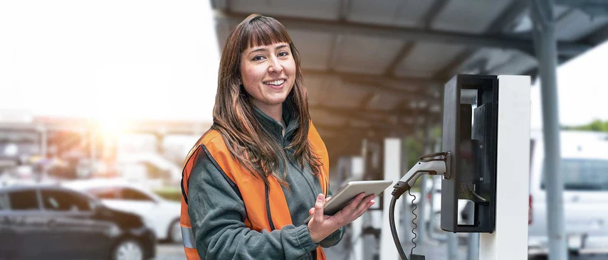 Technikerin in Warnweste mit Tablet an Ladestation für Elektrofahrzeuge in Industriehalle.