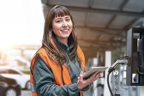 Technikerin in Warnweste mit Tablet an Ladestation für Elektrofahrzeuge in Industriehalle.