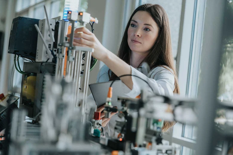 Technikerin arbeitet an technischer Anlage mit elektronischen Bauteilen in hellem Labor. Im Vordergrund weitere Geräte.