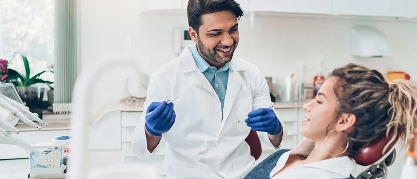 Zahnarzt in weißem Kittel mit blauen Handschuhen behandelt lächelnde Patientin auf Behandlungsstuhl in heller Praxis.