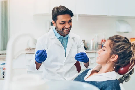 Zahnarzt in weißem Kittel mit blauen Handschuhen behandelt lächelnde Patientin auf Behandlungsstuhl in heller Praxis.