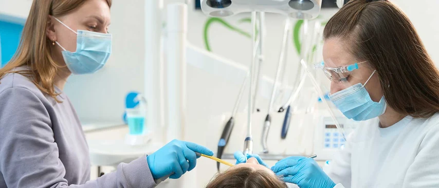 Zahnmedizinische Fachangestellte und Zahnärztin mit Masken und Handschuhen behandeln Patientin auf Behandlungsstuhl.