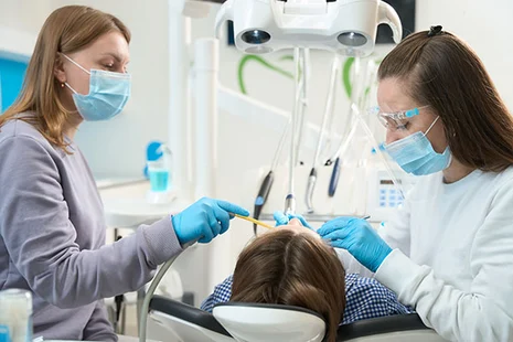 Zahnmedizinische Fachangestellte und Zahnärztin mit Masken und Handschuhen behandeln Patientin auf Behandlungsstuhl.