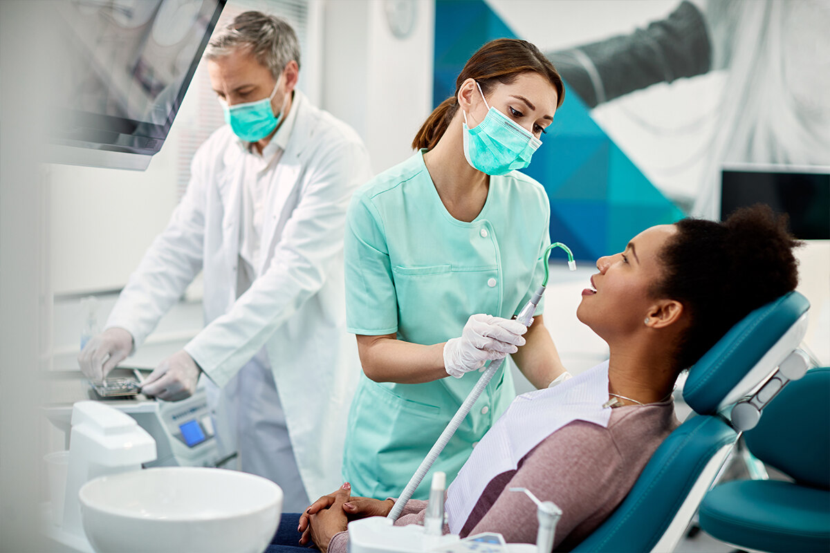 Behandlungszimmer beim Zahnarzt. Patientin sitzt auf Stuhl und wird von Zahnmedizinischer Fachangestellten behandelt. Im Hintergrund ist der Zahnarzt zu sehen.