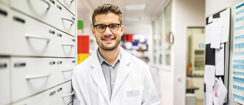 Apotheker in weißem Kittel lächelt in moderner Apotheke mit Schubladen und Regalen im Hintergrund. Experte für Arzneimittelversorgung, Beratung und Qualitätssicherung bei Medikamenten.