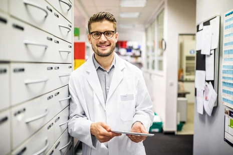 Apotheker in weißem Kittel lächelt in moderner Apotheke mit Schubladen und Regalen im Hintergrund. Experte für Arzneimittelversorgung, Beratung und Qualitätssicherung bei Medikamenten.