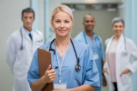 Medizinisches Team im Klinikflur: Pflegekraft mit Stethoskop und Klemmbrett im Fokus, Ärzte im Hintergrund. Medizinische Fachangestellte assistieren bei Behandlungen, betreuen Patienten und koordinieren Praxisabläufe.