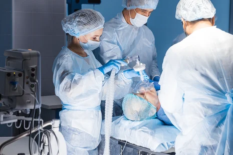 OP-Team bei Beatmungsmanagement: Fachkräfte in Schutzkleidung bereiten Beatmungsmaske und -schlauch für narkotisierten Patienten vor. ATAs überwachen Vitalfunktionen und sichern Atemwege während Operationen.