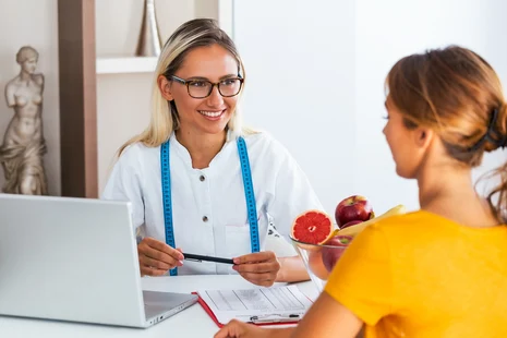 Diätassistentin berät Patientin mit Maßband und frischem Obst am Laptop. Diätassistenten erstellen individuelle Ernährungspläne, berechnen Nährstoffbedarf und schulen bei therapeutischer Ernährungsumstellung.