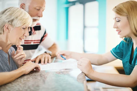 Medizinische Fachangestellte (MFA) berät Seniorenpaar in Arztpraxis: Freundliche Patientenberatung mit Dokumenten am Tisch. MFA koordinieren Termine, empfangen Patienten und verwalten Praxisabläufe.