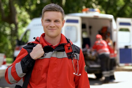 Ein junger Notfallsanitäter laechelt zufrieden in die Kamera, während seine Kollegen in Hintergrund den Krankenwagen bestuecken.