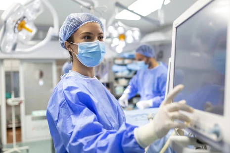 Operationstechnische Assistentin (OTA) im OP mit Haube und Maske bedient Monitor. OTA bereiten Instrumente vor, assistieren bei Operationen, überwachen medizintechnische Geräte und sorgen für sterile Bedingungen im OP-Saal.