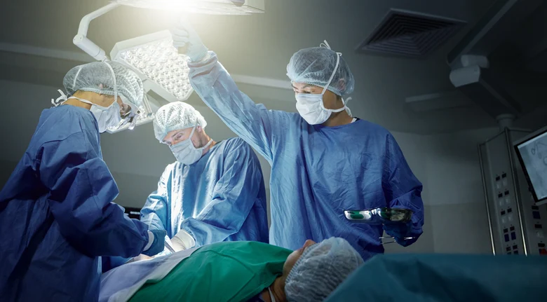 Chirurgisches OP-Team in Schutzkleidung mit Hauben und Mundschutz bei Operation unter OP-Lampe. Teamarbeit unter strengen Hygienestandards: sterile Kittel, Handschuhe und aseptische Techniken schützen Patienten vor Infektionen.