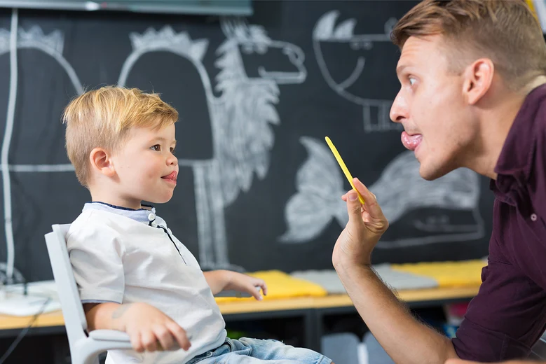 Logopäde übt mit Kind Lautbildung und Zungenstellung vor Anatomietafel. Logopäden behandeln Sprach-, Sprech-, Stimm- und Schluckstörungen bei Kindern und Erwachsenen durch gezielte Übungen zur Verbesserung der kommunikativen Fähigkeiten.