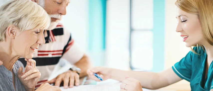 MFA berät Seniorenpaar am Empfangstresen mit Unterlagen. Patientenbetreuung: Medizinische Fachangestellte informieren zu Behandlungen, klären organisatorische Fragen und sind erste Ansprechpartner für Patienten in Arztpraxen und Gesundheitseinrichtungen.